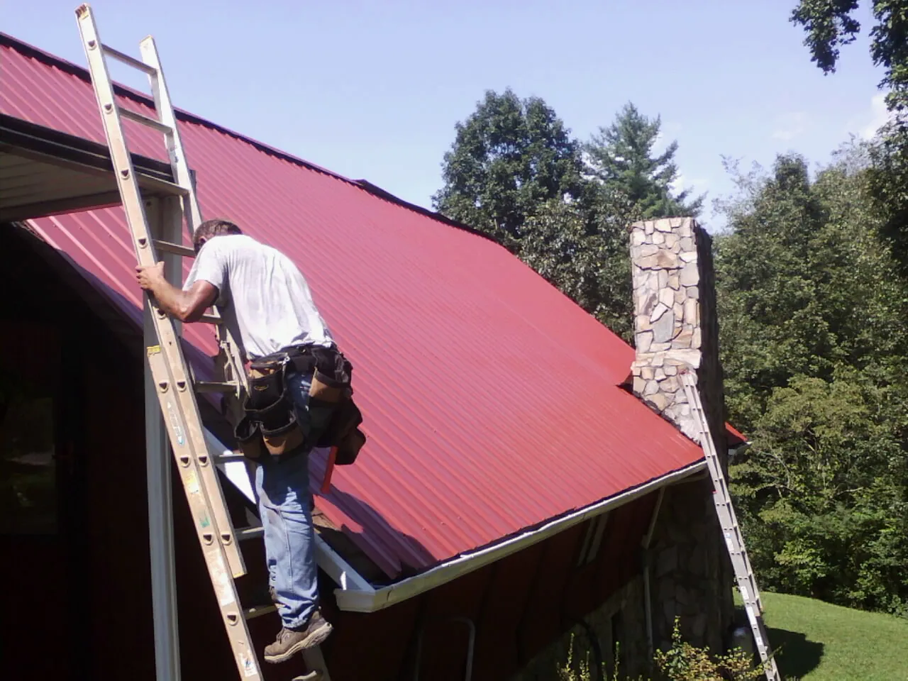 Roofer on a ladder beside a red metal roof and stone chimney — Soffit & Fascia Repair by Roofing Brothers in Bellows Falls